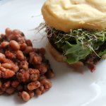 White plate with a portobello mushroom burger topped with fresh greens, avocado mash and baked beans