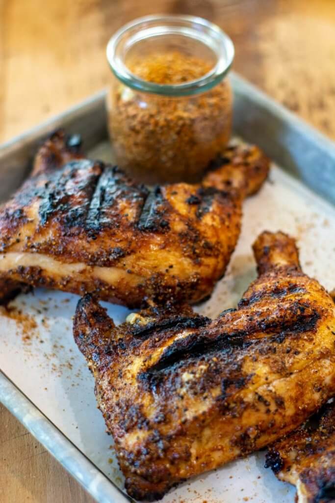 Grilled chicken legs on a baking sheet with the homemade dry rub in a decorative jar.