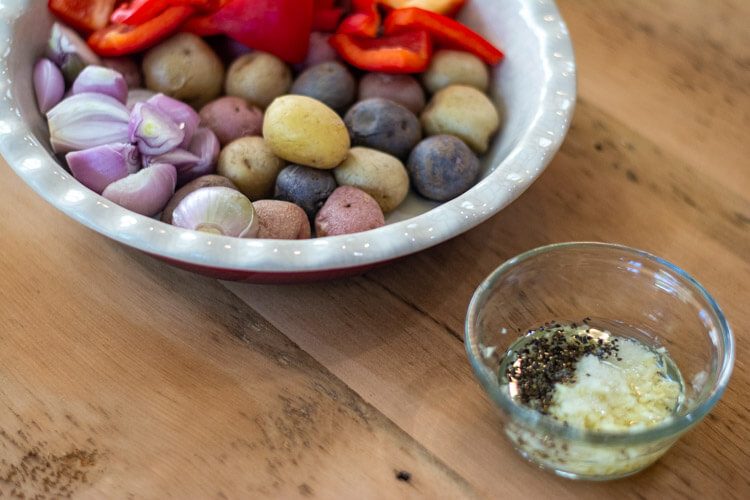 chopped red peppers, shallots and baby potatoes in a dish with a small glass bowl of herbs garlic and oil