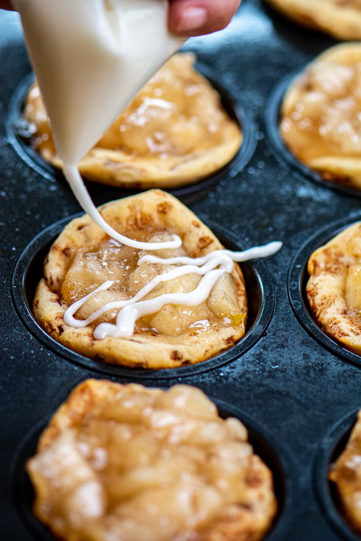 drizzling icing on the apple pie filled cinnamon roll cups in a muffin tin.
