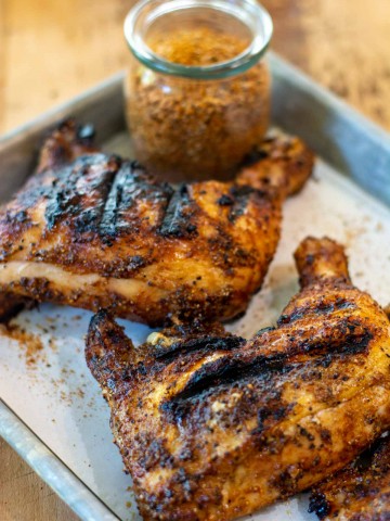 two grilled leg quarters on a lined sheet pan with a decorative jar of BBQ rub.