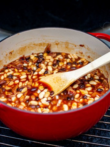 dutch oven on the grill grate with a wooden spoon stirring the grilled baked beans.