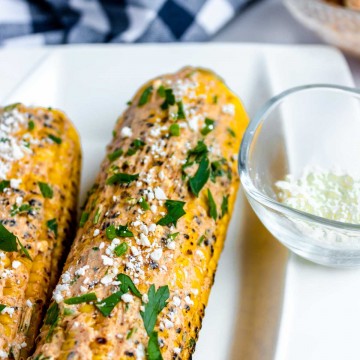 two Mexican Street Corn on a white plate and covered with the mixture and topped with cotija cheese.