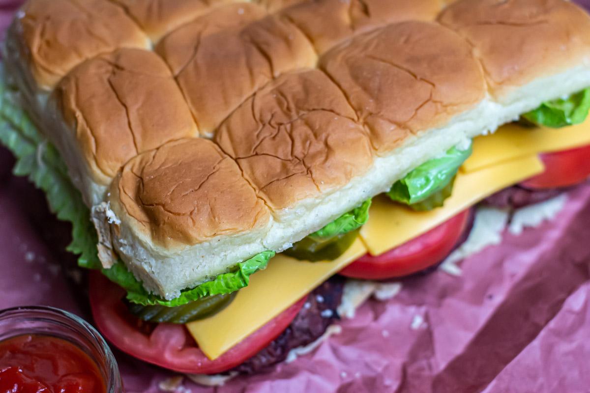 sheet of hawaiian rolls over a platter of hamburger, tomato, pickles, cheese and lettuce.