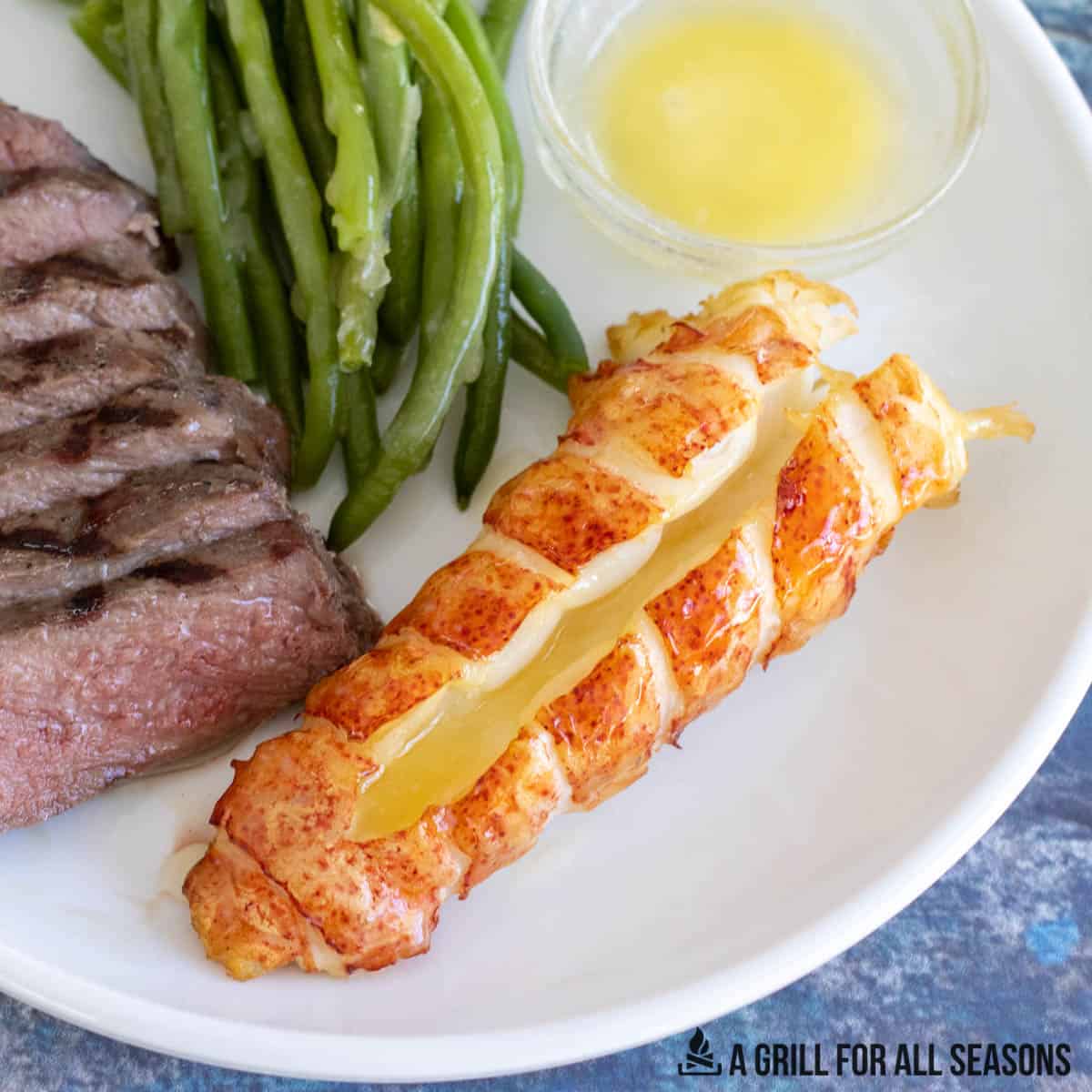 smoked lobster tail with butter on a white plate with green beans and a steak.