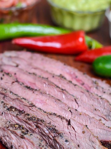 sliced smoked flank steak on a cutting board with peppers, guacamole and pico de gallo.