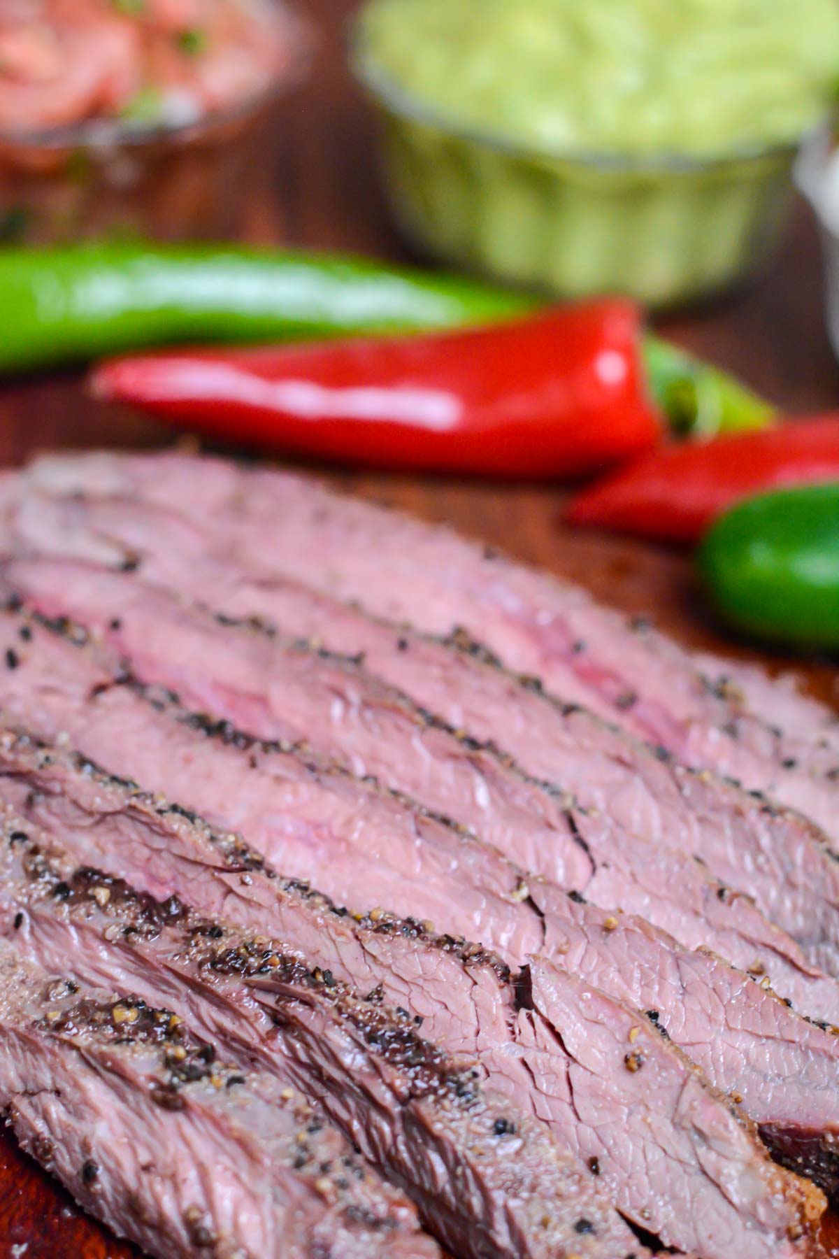 sliced smoked flank steak on a cutting board with peppers, guacamole and pico de gallo.