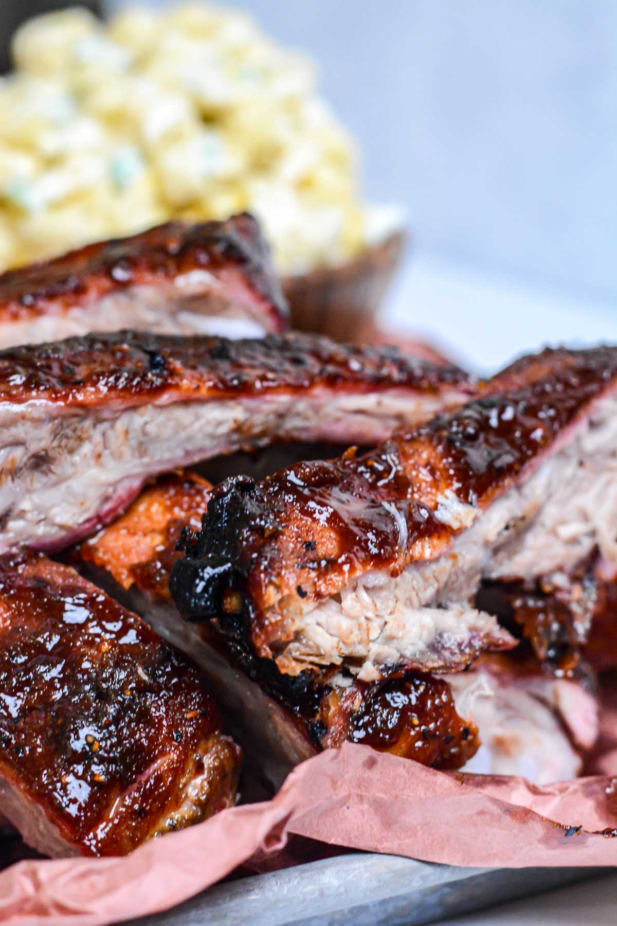 small sheet pan with pink butcher paper and piled with grilled St. Louis Ribs and a bowl of macaroni salad in the background.
