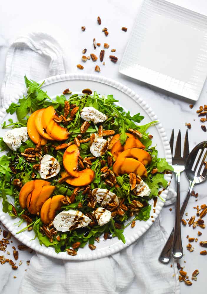 plate loaded with a salad, topped with burrata and peaches and sprinkled with candied nuts.