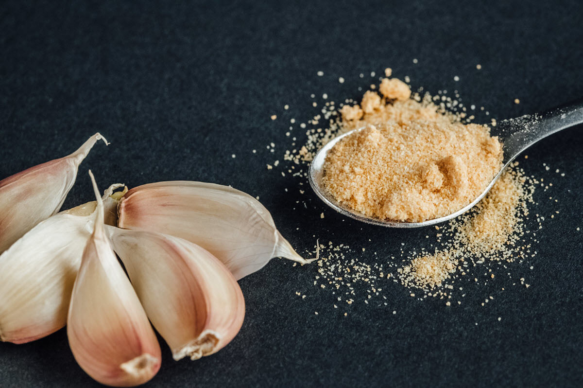 Small spoon of garlic powder and garlic cloves on black table.