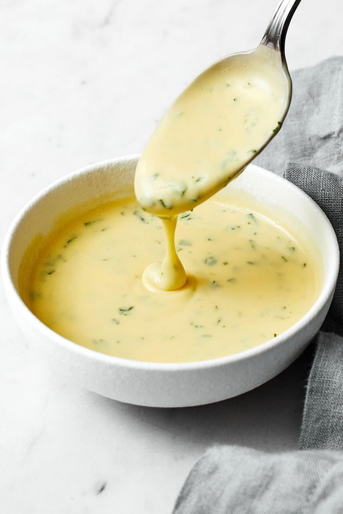 Homemade bearnaise sauce being poured from spoon in white bowl.