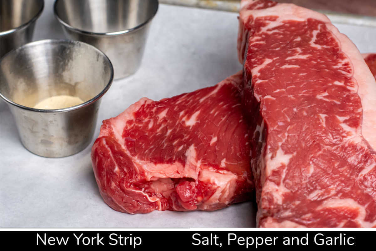 ingredient photo showing two New York Strip steaks and seasoning on a lined sheet pan with labels.