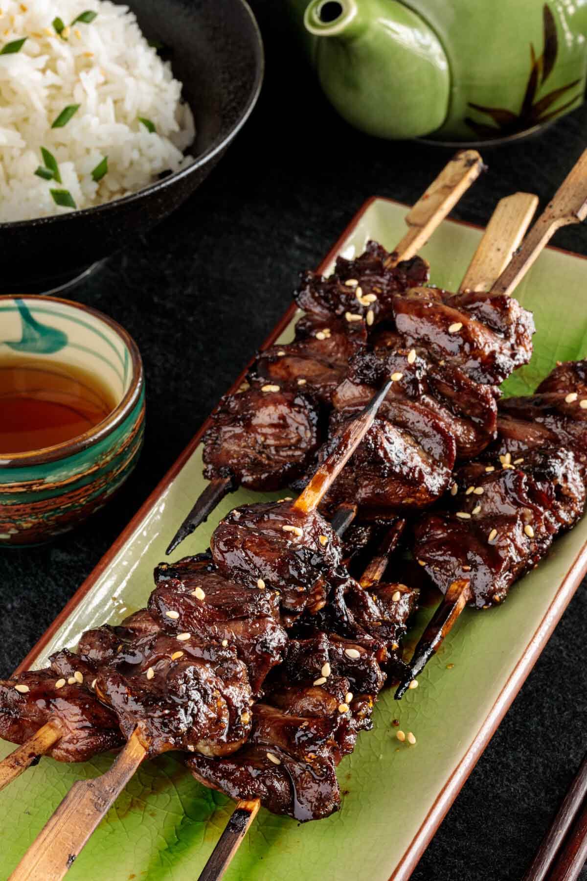 several grilled chicken heart skewers on a platter with rice and tea in the background